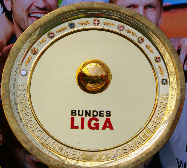 Goldener Schild mit "Bundesliga"-Text in Händen eines Mannes, mit einem Banner im Hintergrund mit drei Personen.