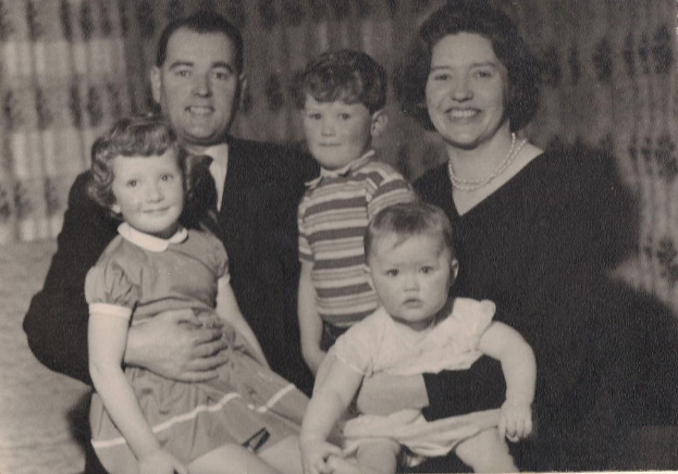 Eine Familie von fünf Personen, darunter ein Mann in Anzug und Krawatte links und eine Frau rechts sowie drei strahlende Kinder, die gemeinsam posieren.
