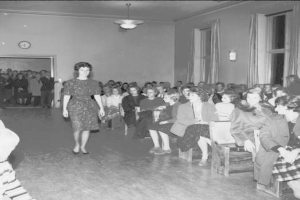 Eine Frau in einem schwarzen und weißen Kleid läuft über den Laufsteg während einer Modenschau für Frauenrechte, mit einer Menge, die auf Stühlen sitzt und einer Uhr und Lichtern an den umliegenden Wänden.