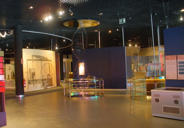 Ein großer Raum mit verschiedenen Geräten, darunter ein metallisches Objekt auf dem Boden, eine Säule, ein Feuerlöscher, Texttafeln, Metallstangen, ein Schild, Deckenleuchten und eine Vitrine.