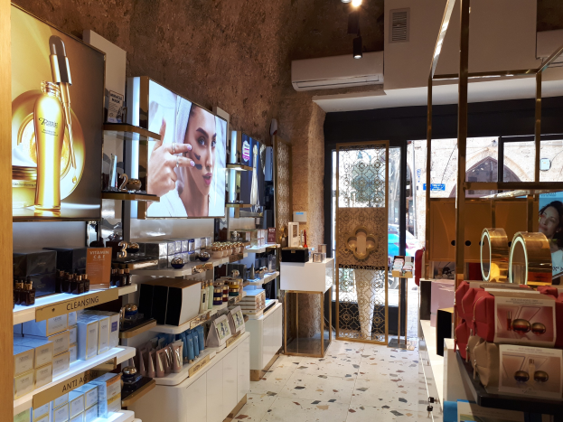 Innenansicht eines Kosmetikgeschäfts mit verschiedenen Produkten auf Gestellen, Wandbildschirmen, Deckenleuchten, einer Klimaanlage und Glastüren, die eine Person, ein Auto und ein Gebäude draußen zeigen.