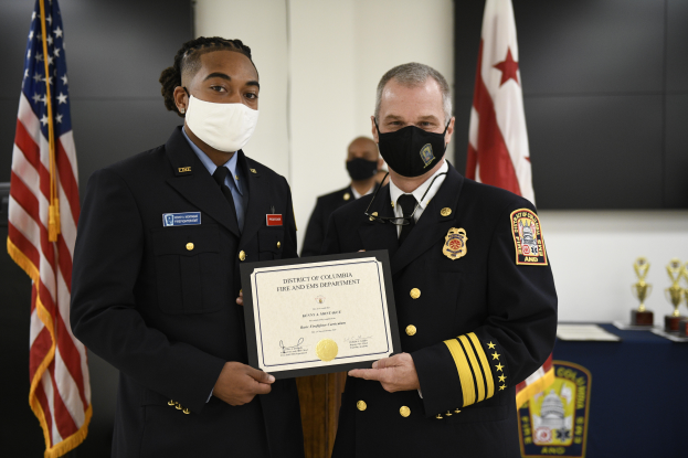 Zwei Männer in Uniform, die Masken tragen und ein Zertifikat halten, stehen nebeneinander vor einem Tisch, der mit einer blauen Decke bedeckt ist und mit Schilden und anderen Gegenständen geschmückt ist, mit Flaggen und einer Wand im Hintergrund.