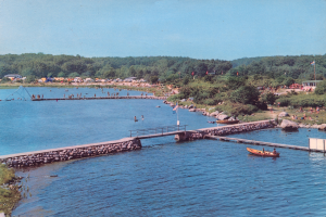 Ein altes Foto eines Sees mit einem Boot, einer Brücke, Flaggen, Felsen, Bäumen, Gebäuden und einem Himmel im Hintergrund, das viele Menschen beim Schwimmen zeigt und Text auf dem Bild geschrieben hat.