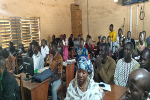 Eine Gruppe von Menschen sitzt an Tischen in einem Klassenzimmer mit Monitoren, Taschen, Büchern, Papieren und anderen Gegenständen auf den Tischen, einem Fenster auf der linken Seite und Kartons und einem Fernseher an der Wand im Hintergrund, die an einer Technologie-Schulung teilnehmen.