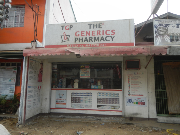 Ein Apotheken-Schild mit der Aufschrift "The Generics Pharmacy" an einem Gebäude in einer städtischen Umgebung mit sichtbarer Infrastruktur und Fahrzeugen unter einem klaren Himmel.