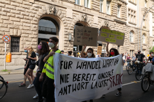 Eine Gruppe von Menschen marschiert auf einer Straße bei einer Demonstration, einige halten Schilder und andere fahren Fahrräder, im Hintergrund ist ein Gebäude mit Fenstern, Bögen, Säulen und Skulpturen zu sehen.