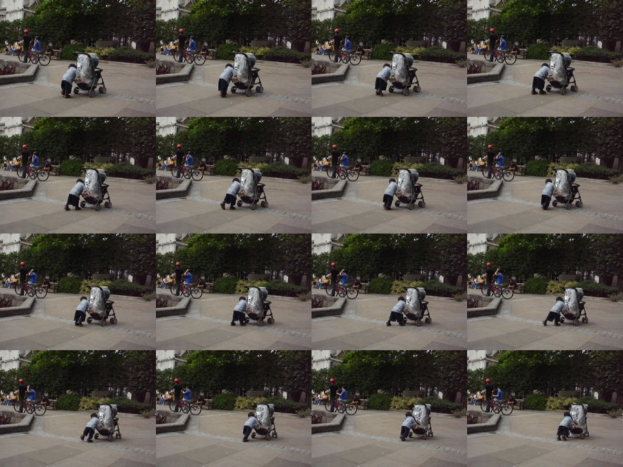 Ein Kind geht auf einer Straße und zieht einen Karren, mit zwei Personen, die Fahrräder halten, links, Bäumen im Hintergrund und einem Gebäude links.