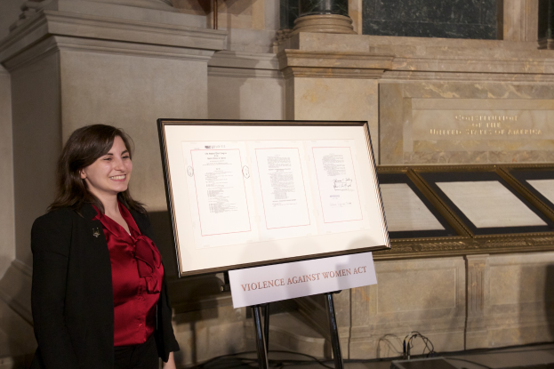Eine Frau in roter Bluse und schwarzer Jacke lächelt vor einem Schild, auf dem "Gesetz gegen Gewalt gegen Frauen" steht, mit einer Wand und Säulen im Hintergrund.