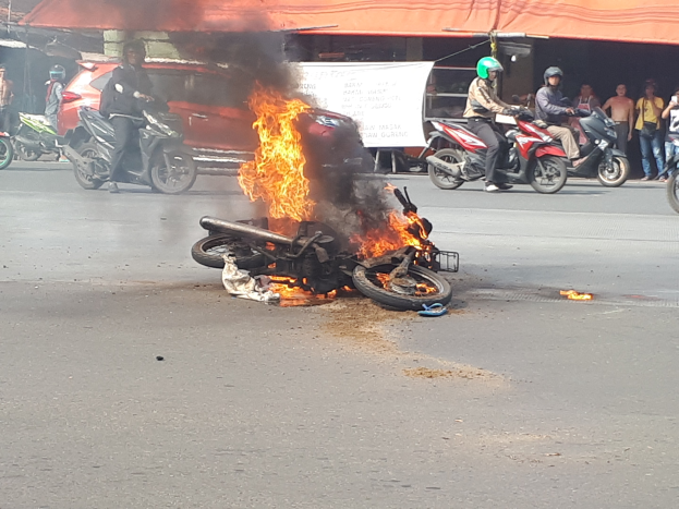 Ein Motorrad steht in Flammen am Straßenrand, mit einem Auto und einem Transporter im Hintergrund, umgeben von Menschen, einige tragen Helme, und eine Fahne mit Text im Hintergrund, sowie ein Zelt.