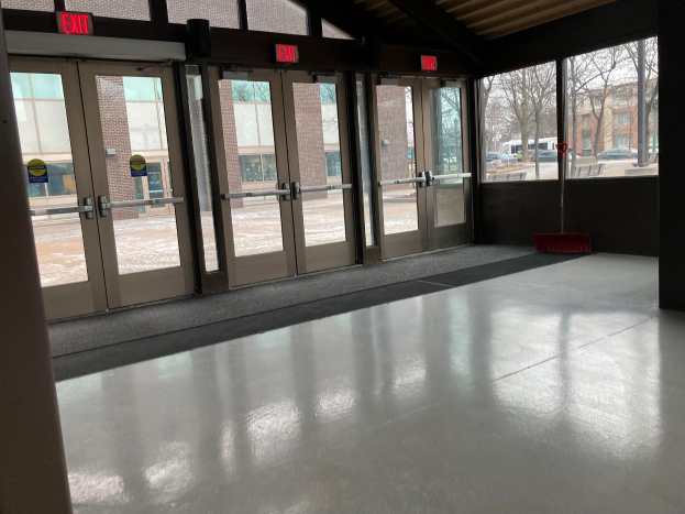 Großer Raum mit hohen Decken, mehreren Fenstern und Türen, mit einem Besen auf dem Boden, Schildern mit Text, einem sichtbaren Dach, umliegenden Bäumen, Fahrzeugen auf dem Boden, benachbarten Gebäuden mit Fenstern und einem bewölkten Himmel, was auf einen gewerblichen Bodenreinigungsservice hinweist.