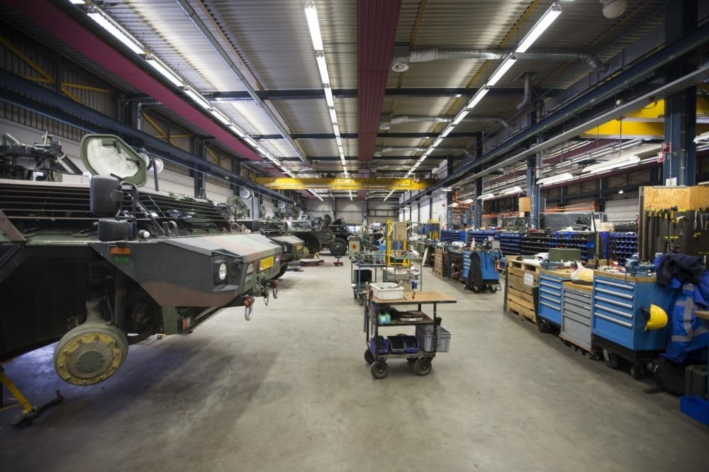 Großes Lager mit Maschinen, Fahrzeugen auf der linken Seite, Wagen mit Gegenständen, Holzboxen und Deckenbeleuchtung.