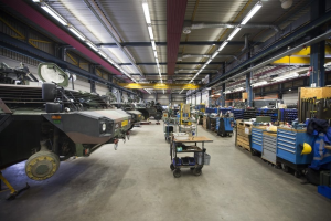 Großes Lager mit Maschinen, Fahrzeugen auf der linken Seite, Wagen mit Gegenständen, Holzboxen und Deckenbeleuchtung.