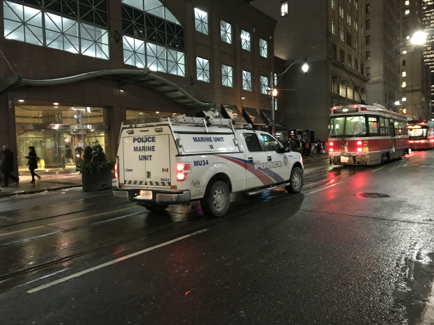 Ein Polizei-Lkw steht nachts am Straßenrand einer Stadt, mit Fahrzeugen auf der Straße, Fußgängern auf dem Gehweg, Topfpflanzen, Straßenlaternen, Gebäuden mit Fenstern und einer bewölkten Himmel.