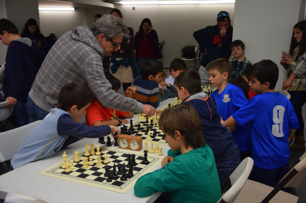 Kinder spielen Schach in einem Wettbewerb, während Erwachsene von hinten und den Seiten zuschauen.