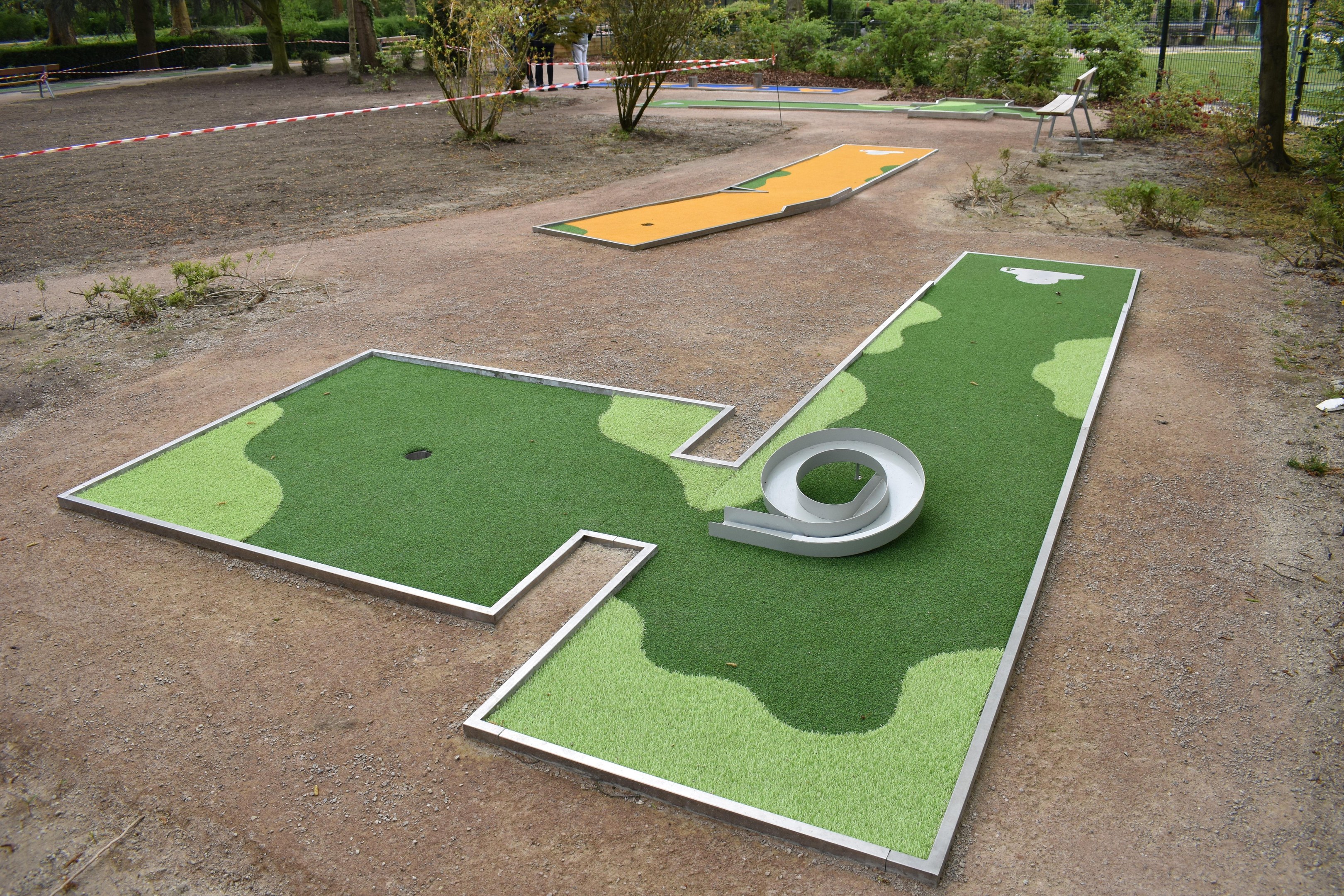 Minigolfplatz in einem Park mit Grünflächen, Menschen, Gebäuden und einem klaren Himmel.