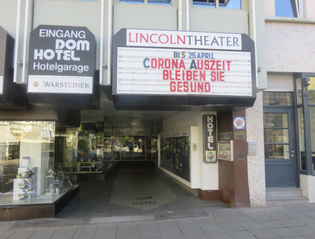 Außenansicht des Lincoln Theaters in Berlin, Deutschland, mit Glasfenstern und -türen sowie einer Tafel mit Text und einem Inneren, das eine belebte Stadtlandschaft suggeriert.