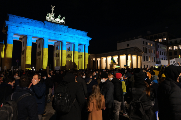Eine Menschenmenge steht vor dem Brandenburger Tor in Berlin, Deutschland, viele tragen Mützen und Taschen, einige halten Schilder, mit den Säulen und Statuen des Tors im Hintergrund, Gebäuden mit Fenstern im Hintergrund, unter einem dunklen Himmel.