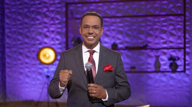 Stephen Colbert, Moderator von 'The Daily Show with Trevor Noah', steht auf einer Bühne und hält ein Mikrofon, trägt einen Anzug, eine Krawatte und eine Brille, mit einer Lampe und einem Ständer im Hintergrund.