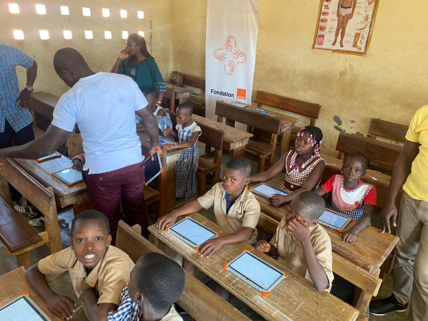 Eine Gruppe von Kindern sitzt an Tischen in einem Klassenzimmer und verwendet Tablets, während einige Menschen um sie herumstehen und ein Banner und ein Plakat im Hintergrund zu sehen sind.