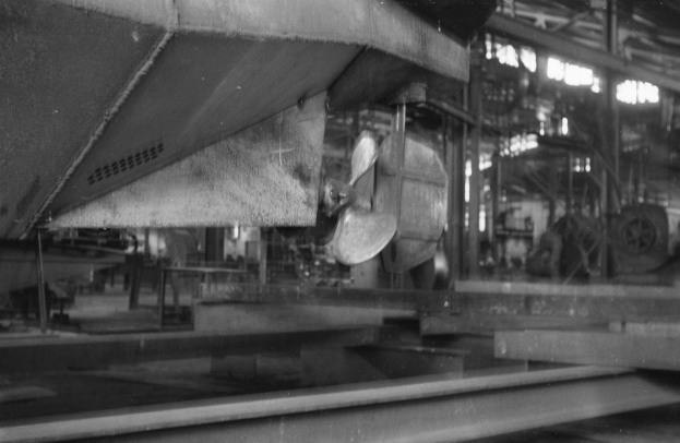 Ein Schwarz-Weiß-Foto eines Boots in der Fertigung in einer Fabrik, umgeben von metallischen Gegenständen, Pfählen und Lichtern.