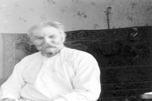 Ein alter Mann mit Schnurrbart sitzt in einem Stuhl vor einer Wand, trägt ein weißes Hemd und hat einen ernsten Ausdruck im Gesicht, auf einem Schwarz-Weiß-Foto.