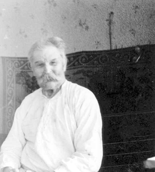 Ein alter Mann mit Schnurrbart sitzt in einem Stuhl vor einer Wand, trägt ein weißes Hemd und hat einen ernsten Ausdruck im Gesicht, auf einem Schwarz-Weiß-Foto.