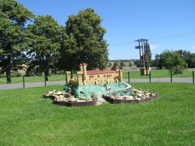 Miniatur-Golfplatz mit Hindernissen auf einem Rasenfeld, umgeben von einem Maschendrahtzaun, umgeben von Bäumen, Strommasten und einem bewölkten Himmel.