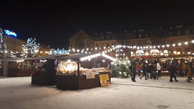 Ein belebter Weihnachtsmarkt an einem verschneiten Abend mit Menschen, Buden mit Lichtern, Pflanzen, Bäumen, Gebäuden und Schildern.
