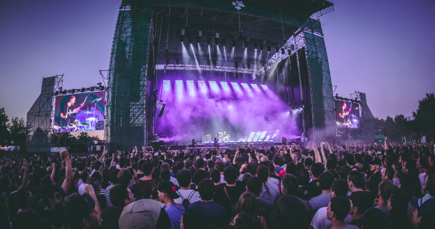 Eine große Menschenmenge auf einem Musikfestival mit einer Bühne in der Mitte, auf der Musiker Instrumente spielen, beleuchtet von Scheinwerfern und umgeben von Bildschirmen, mit einem Gebäude, Bäumen und einem klaren blauen Himmel im Hintergrund.