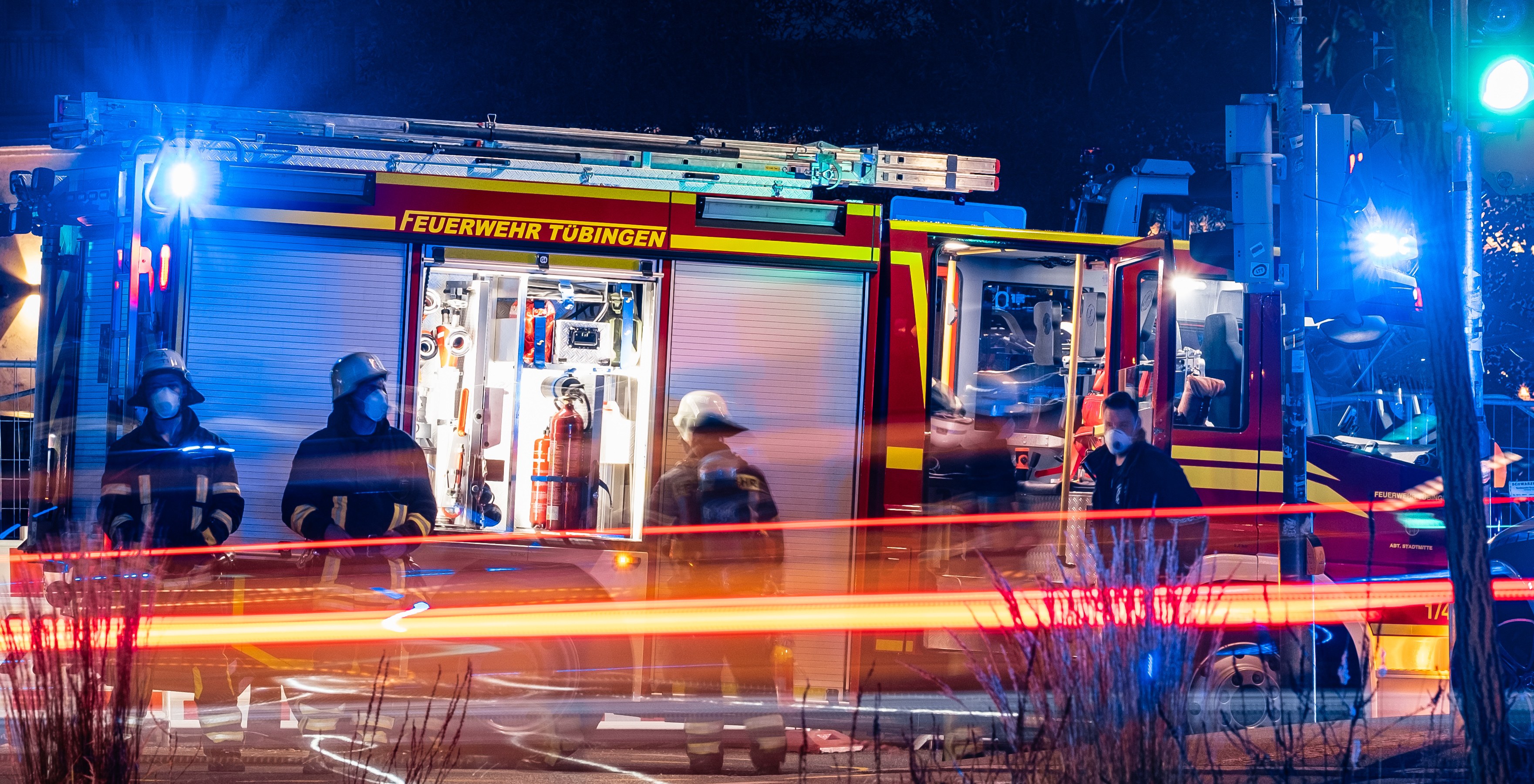 Feuerwehrleute in Schutzausrüstung vor einem beleuchteten Feuerwehrauto bei Nacht, mit Pflanzen im Vordergrund, einem Verkehrszeichen auf der rechten Seite und Bäumen im Hintergrund.