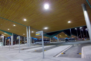 Bushaltestelle nachts mit Deckenbeleuchtung, Fahrzeuge auf der Straße, Beschilderung, Menschen und beleuchtete Gebäude im Hintergrund.