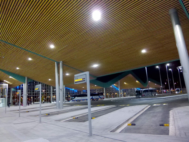 Bushaltestelle nachts mit Deckenbeleuchtung, Fahrzeuge auf der Straße, Beschilderung, Menschen und beleuchtete Gebäude im Hintergrund.