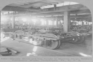 Schwarzes und weißes Foto eines Elektromotors und eines Lastwagens in einer Fabrik mit Maschinen, Säulen und einem Dach, einschließlich Text unten.