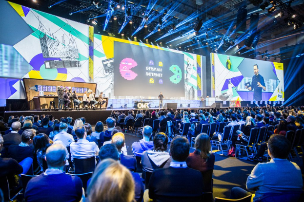 Eine große Menschenmenge sitzt auf Stühlen vor einer Bühne bei der Deutschen Internetkonferenz 2019, mit einem Tisch voller Gegenstände links und Bildschirmen und Lichtern im Hintergrund.
