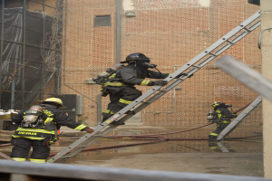Feuerwehrleute in Helmen und Ausrüstung klettern an einer Leiter vor einem Backsteingebäude mit Rohren auf dem Boden und einer Metallstange unten, mit einem weiteren Gebäude und einem Netz im Hintergrund.