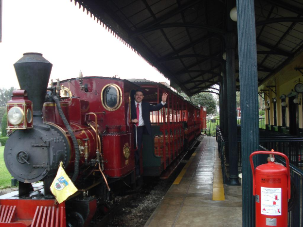 Ein altes rotes Zug mit einem Mann in einem schwarzen Anzug, der an der Tür steht und lächelt, mit einem Bahnhofsgebäude auf der rechten Seite und einem roten Feuerlöscher im Vordergrund.