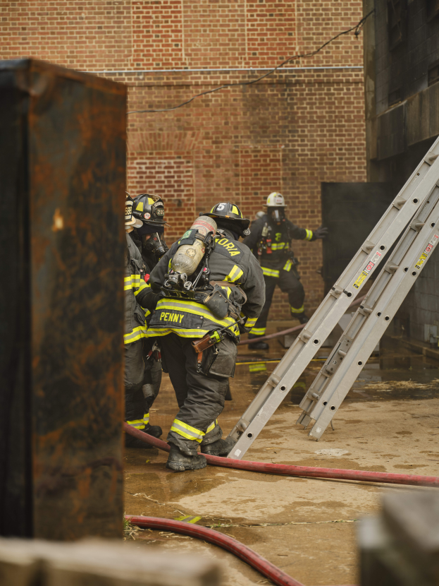 Feuerwehrleute mit Helmen und Handschuhen arbeiten daran, ein Gebäudefeuer zu löschen, mit einer Leiter auf der rechten Seite und einem Metallobjekt auf der linken Seite, vor einem Backsteinwällenhintergrund.