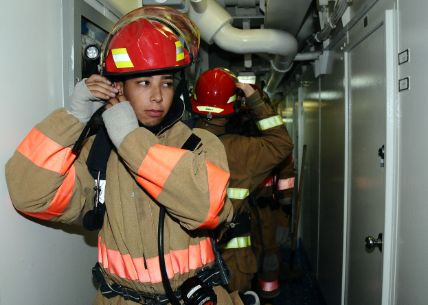 Eine Gruppe von Feuerwehrleuten in Uniform, die zusammen in einem Trainingsraum mit Rohren und Equipment im Hintergrund stehen.