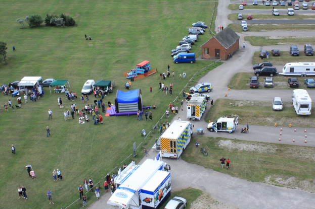 Eine Luftaufnahme eines Campingplatzes mit vielen geparkten Fahrzeugen, einigen Menschen und einer zentralen Hütte sowie einigen Bäumen auf der linken Seite.