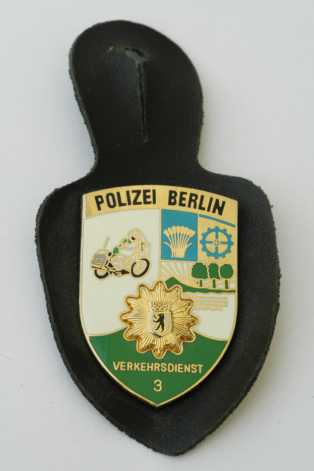 Ein schildförmiges Polizeibadged mit der Aufschrift "Polizei Berlin" in fetter schwarzer Schrift auf einem blauen Hintergrund mit einem weißen Stern und Rahmen, das auf einer Oberfläche liegt.