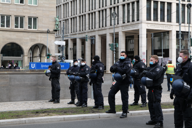 Eine Gruppe von Polizisten in schwarzen Uniformen und Masken steht vor einem Gebäude mit Glasfenstern und Säulen in Berlin, Deutschland, einige halten Helme, mit Laternenmasten, Verkehrsampeln, Tafeln mit Text, einer Statue, anderen Menschen und einem grasbewachsenen Boden im Hintergrund.