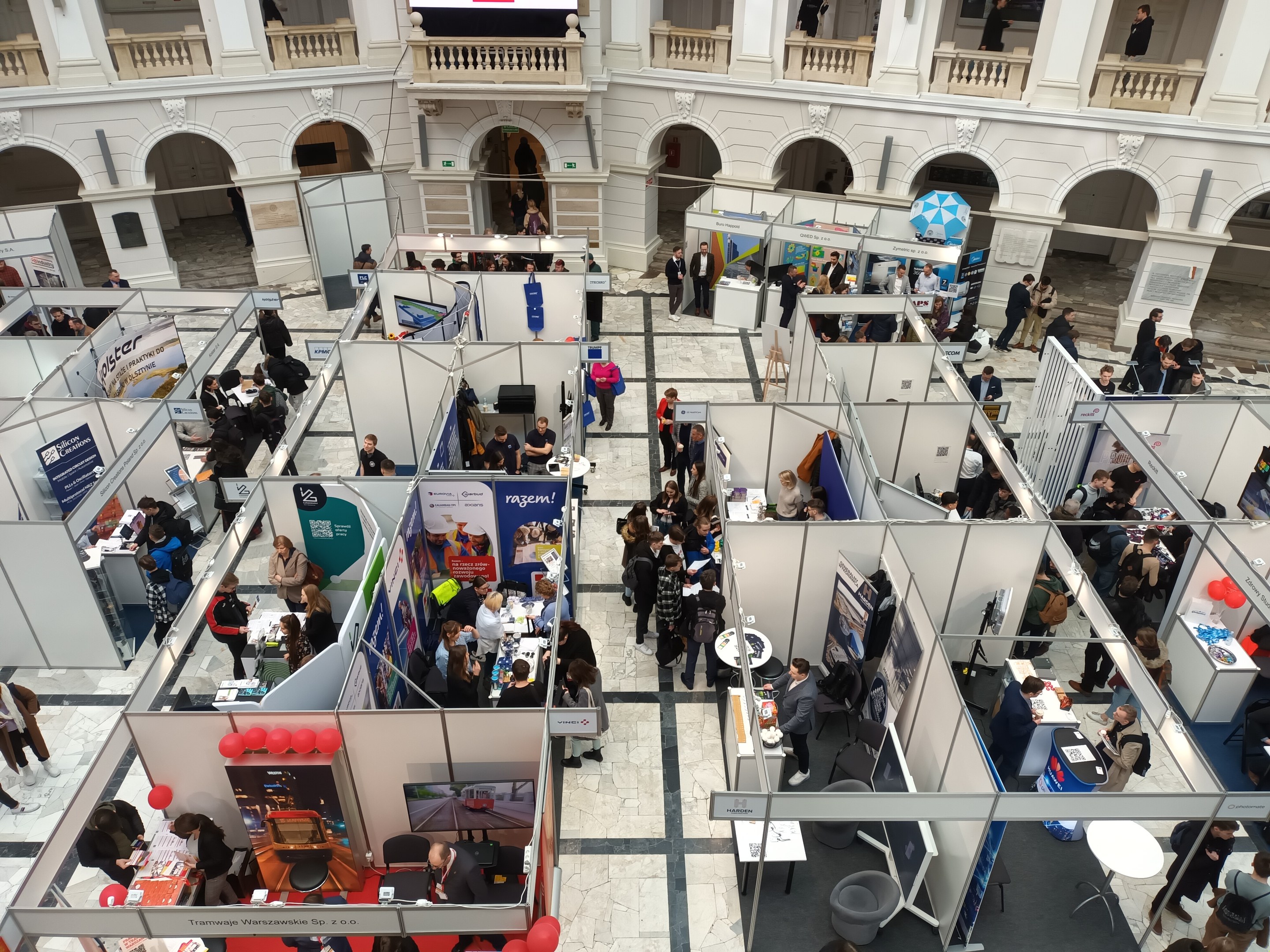 Große Gruppe von Teilnehmern auf einer internationalen Fachmesse mit Ständen, Bannern, Tischen, Stühlen und Ballons in einem Raum mit architektonischen Bögen, Säulen und einem Bildschirm im Hintergrund.