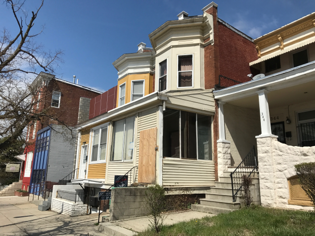 Eine Reihe von baufälligen Häusern an einer Straßenkreuzung, einige verbarrikadiert, mit Glasfenstern, Treppen mit Geländern, Pflanzen, Gras, Bäumen und einem bewölkten Himmel im Hintergrund.
