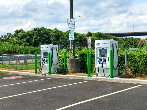 Elektrofahrzeug-Ladestation in einem Parkplatz mit umgebender Infrastruktur, Vegetation und einer Brücke im bewölkten Himmel.