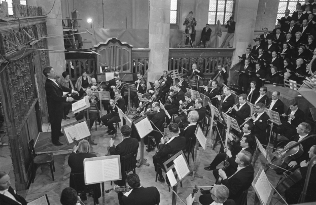 Schwarzes und weißes Bild einer Gruppe von Menschen, die Musikinstrumente in einer Kirche spielen, sitzend und stehend mit Notenpulten, mit Fenstern, Säulen und Lichtern im Hintergrund.