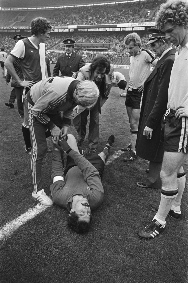 Schwarzes und weißes Bild eines Fußballspielers, der auf dem Boden liegt und von einer Gruppe von Männern umgeben ist, mit einer Menschenmenge im Hintergrund eines Stadions.