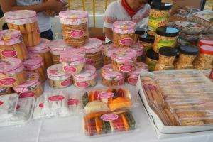 Eine Gruppe von Menschen steht um einen Tisch herum, der mit verschiedenen Lebensmitteln gefüllt ist, darunter Schachteln und Verpackungen, mit einem Geländer und einer Straße im Hintergrund auf einem Food-Festival.