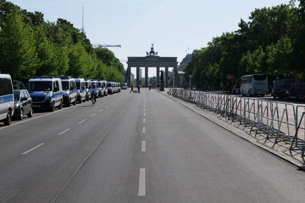 Lange Reihe von Polizeiwagen auf der Straße vor dem Reichstagsgebäude in Berlin, Deutschland, mit Menschen auf Fahrrädern und Barrieren auf der Straße, Bäumen an den Seiten und einem Bogen mit Statuen im Hintergrund.