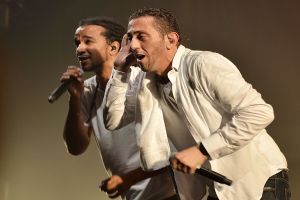 Zwei Männer in legerer Kleidung auf der Bühne mit Mikrofonen, Hintergrund leicht unscharf.