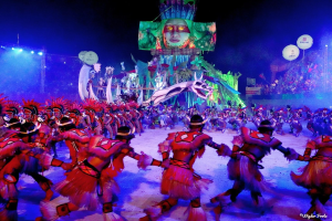 Eine Gruppe von Menschen in bunten Kostümen tanzt auf einer Bühne vor einem sitzenden Publikum, mit Lichtern, Pfählen, Ballons und einem Bildschirm, der eine Person zeigt, unter einem sichtbaren Himmel, bei der Rio-Karneval 2014.